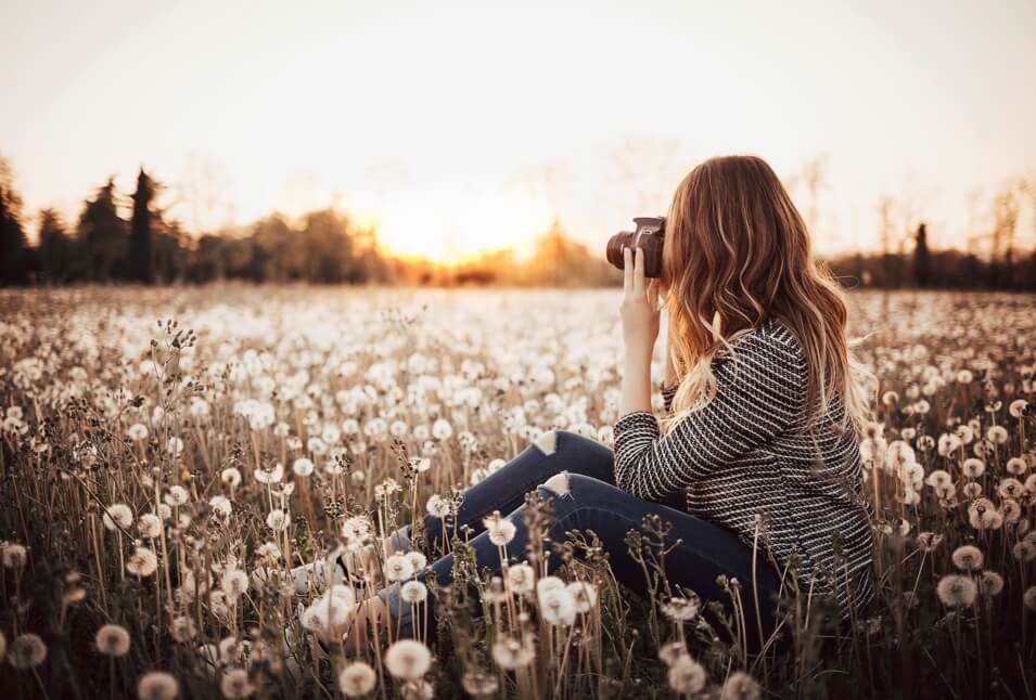 A feminine presenting person sitting in a field of dandelions at golden hour.