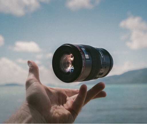 A camera lens suspended in mid-air above an open palm with the ocean, mountains, and a blue sky in the background.